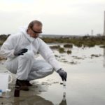 Person in protective gear collecting water samples with tools at an outdoor site for Charles Krull & Son, Inc.