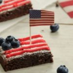 Brownie dessert topped with red frosting and blueberries, featuring an American flag, perfect for 4th of July celebrations.