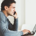 Man on phone in casual sweater at desk with laptop, discussing home maintenance plans for plumbing and heating services.