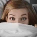Young woman with wide eyes under a light blanket, showing furnace noise concerns for homeowners at Charles Krull & Son.