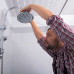 Man adjusting a round perforated showerhead in a bathroom to improve water pressure for plumbing services.