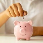 Hand inserting a coin into a pink piggy bank in an indoor setting, illustrating financial responsibility for homeowners.