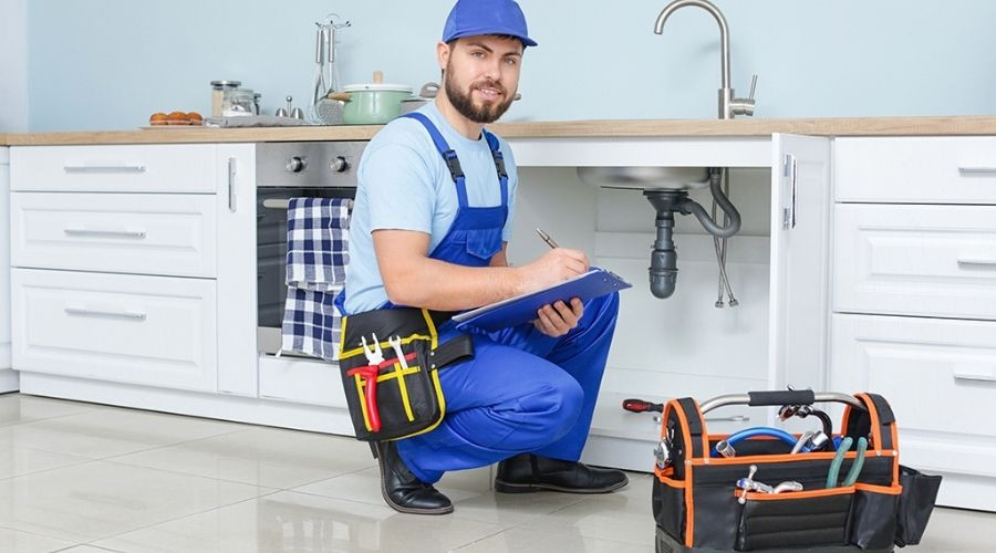Male plumber in blue overalls writing on a clipboard in a modern kitchen with a toolbox. Charles Krull & Son, Inc.