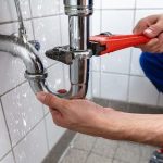 Person's hands holding a red adjustable wrench near a leaking plumbing pipe in a bathroom by Charles Krull & Son, Inc.