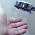 Close-up of a human hand under a chrome showerhead with flowing water, highlighting plumbing maintenance for homes.